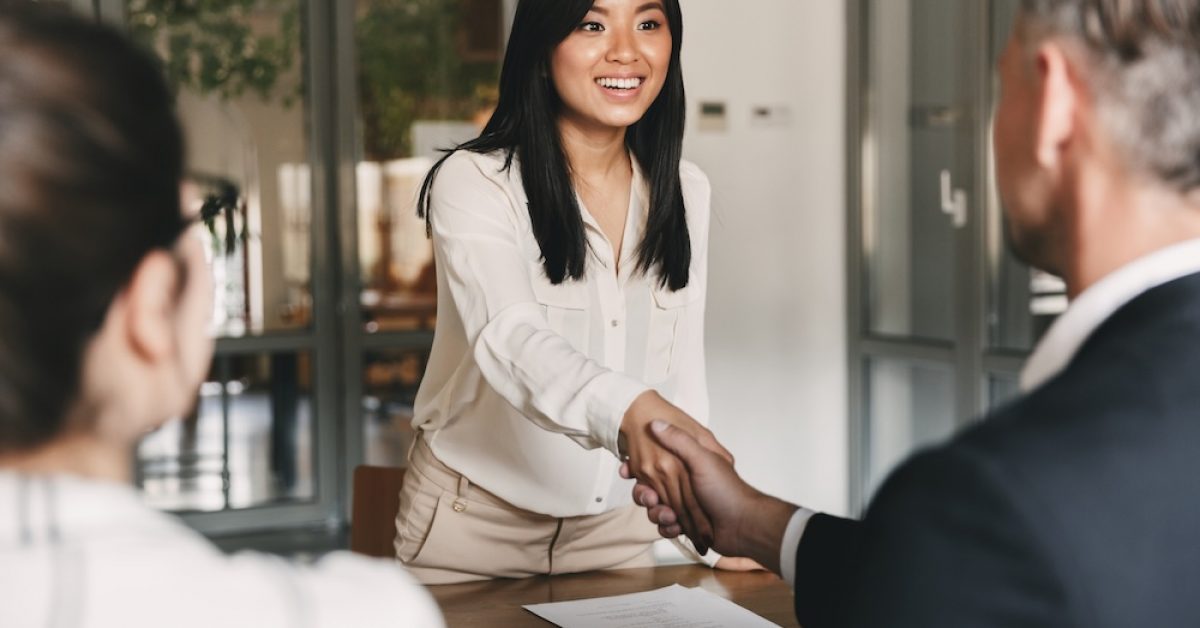 Business, career and placement concept - image from back of two employers sitting in office and shaking hand of young asian woman after successful negotiations or interview